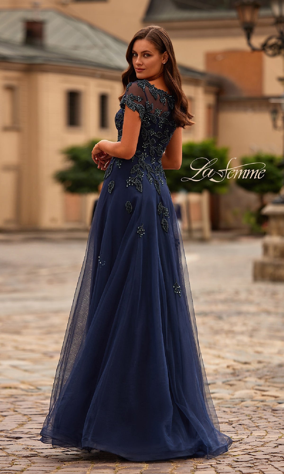 A woman in the La Femme 31692 Short Sleeve Navy Formal Dress stands outdoors on a cobblestone street, looking over her shoulder.